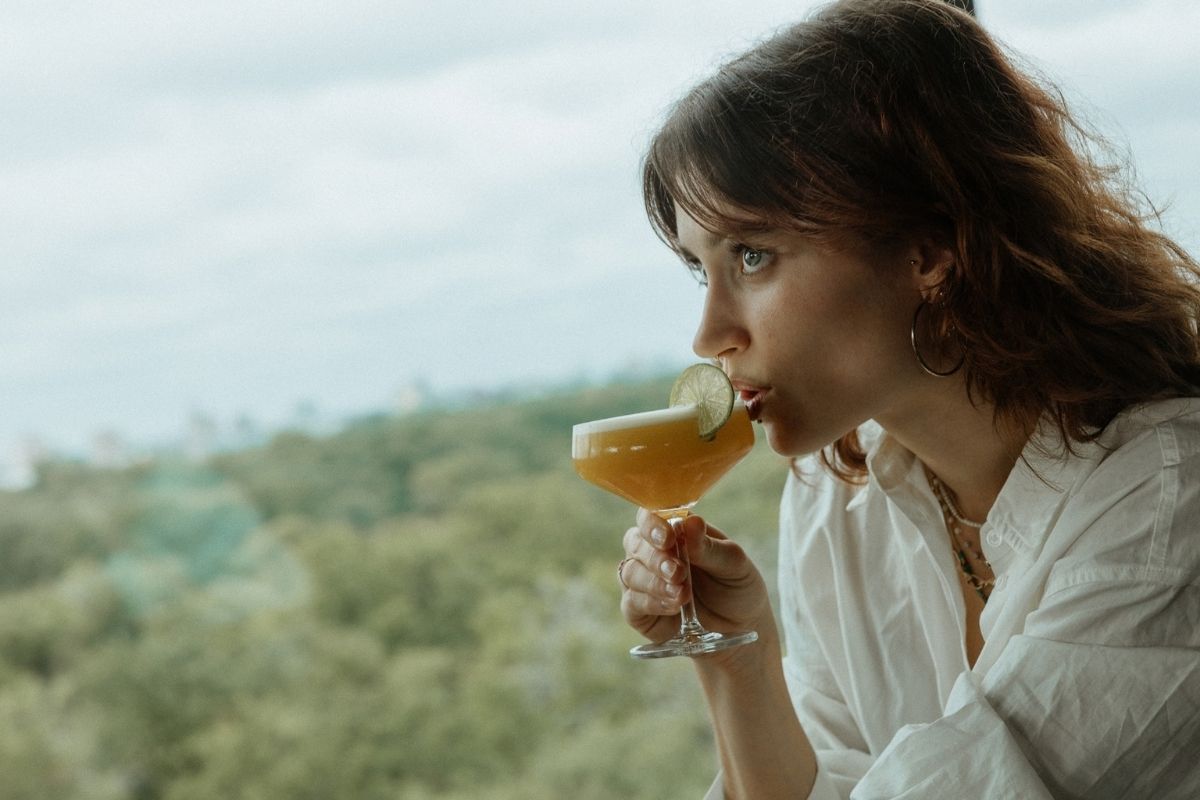 A woman in a white blouse sips an amber cocktail garnished with lime, gazing thoughtfully over a hazy green landscape in a calm.